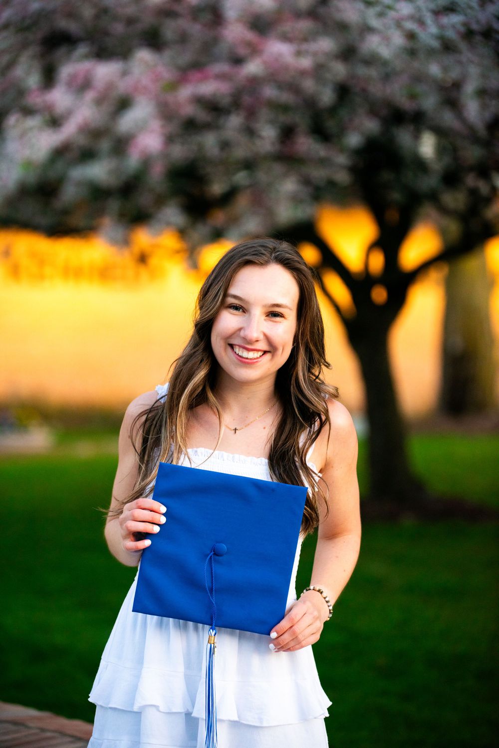 Graduate holding blue diploma cover under blooming cherry tree at sunset.