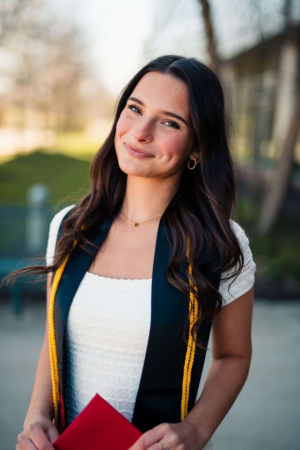 Graduate wearing a navy academic stole with gold trim and white top poses outdoors with their diploma.