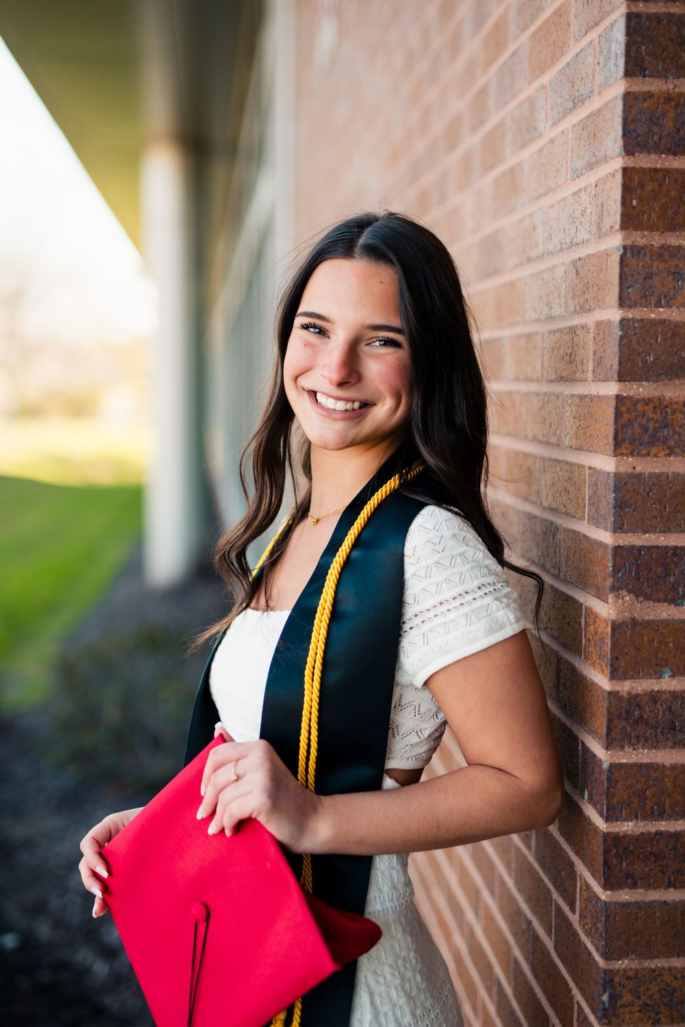 Graduate student in white shirt and dark vest holding red graduation cap poses against brick wall with honor cord.