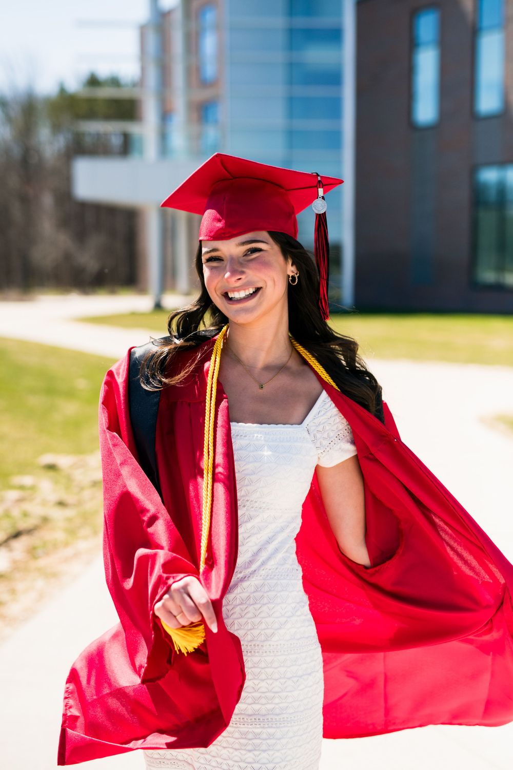 Graduate in bright red cap and gown celebrating outdoors in sunshine wearing white dress on campus.