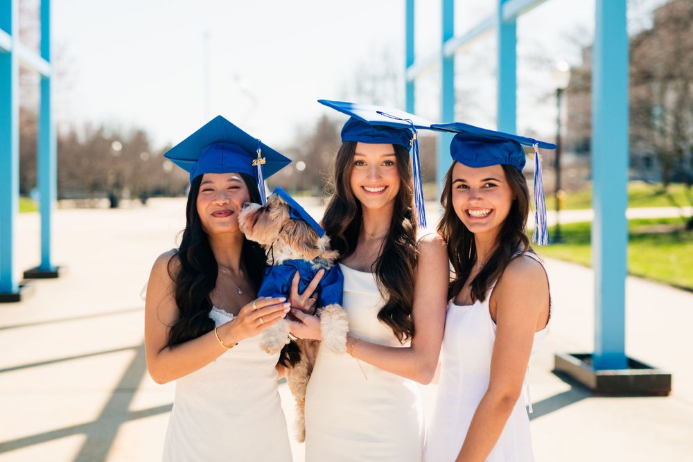 Three college graduates in blue caps and gowns celebrate together outside on a sunny day.
