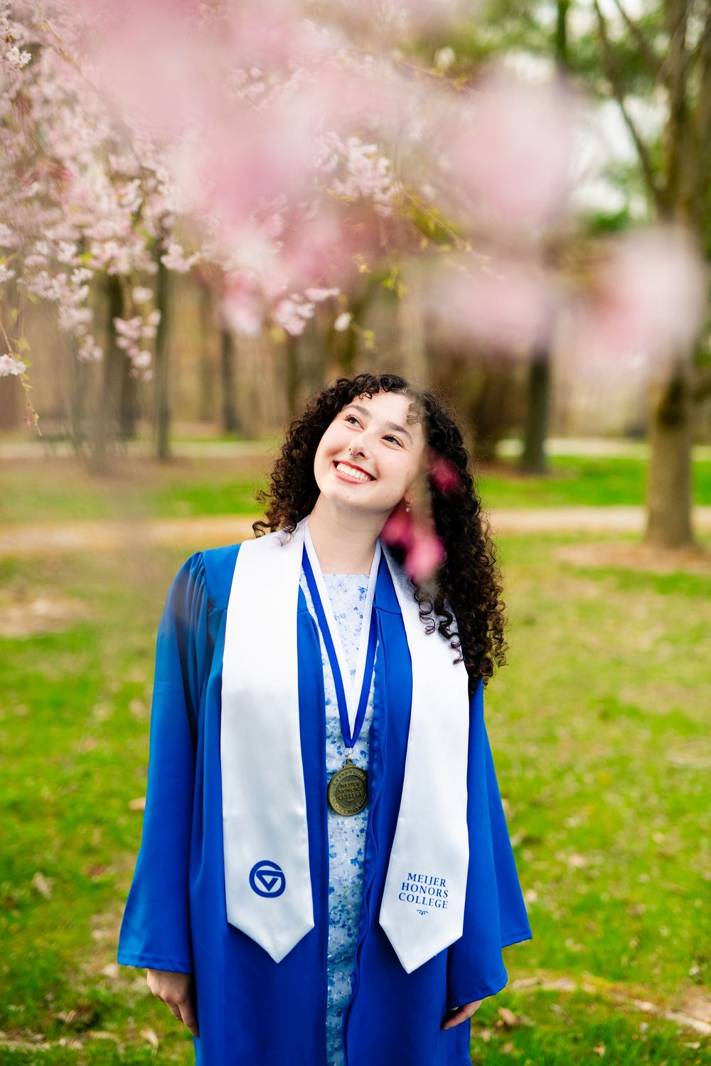 Graduate wearing blue graduation gown and white stole stands smiling under blooming cherry blossoms in spring.