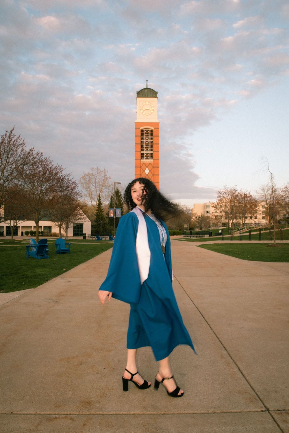 Graduate in blue academic regalia stands on campus path with illuminated clock tower in background at sunset.