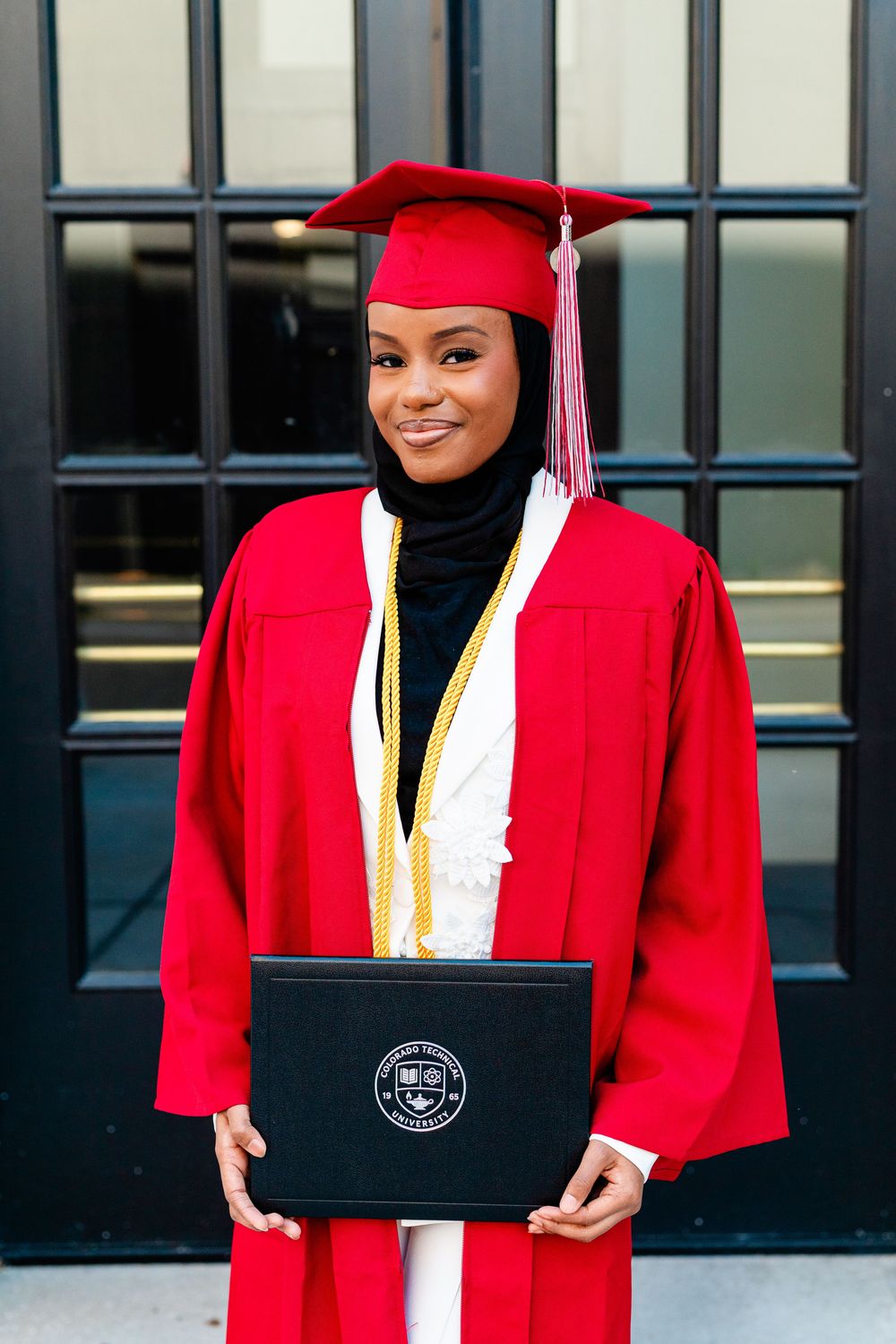 Young woman in red graduation cap and gown holding diploma, wearing hijab with gold honor cords.