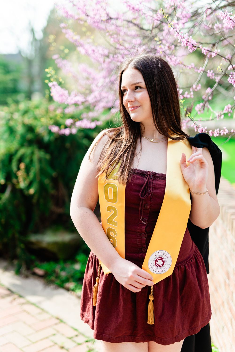 Young woman in maroon dress wearing gold graduation stole with 2025 standing outdoors near pink flowering trees.
