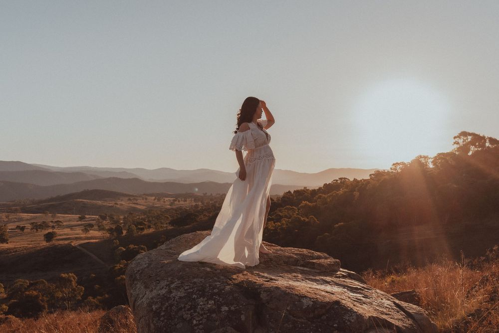 A person in a flowing white dress stands on a rocky outcrop at sunset overlooking a mountainous landscape.