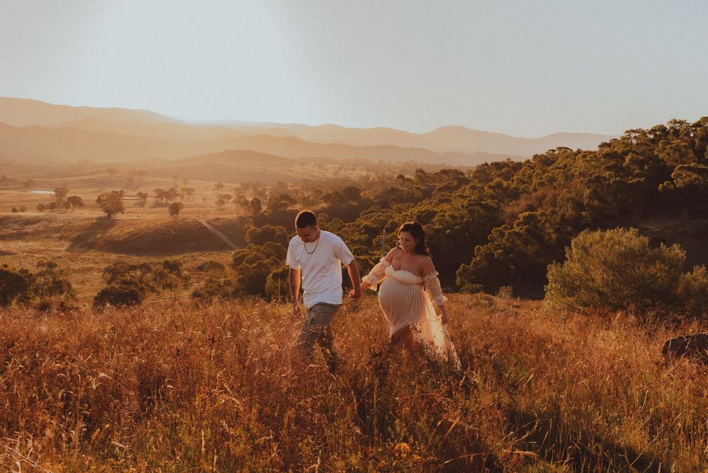 Two figures walk through golden sunlit grass in a mountainous landscape during sunset.