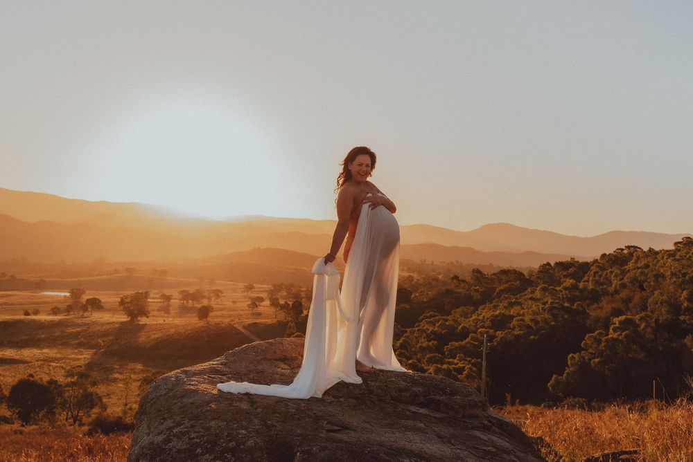 Figure in flowing white dress stands on rocky outcrop overlooking desert landscape at sunset.
