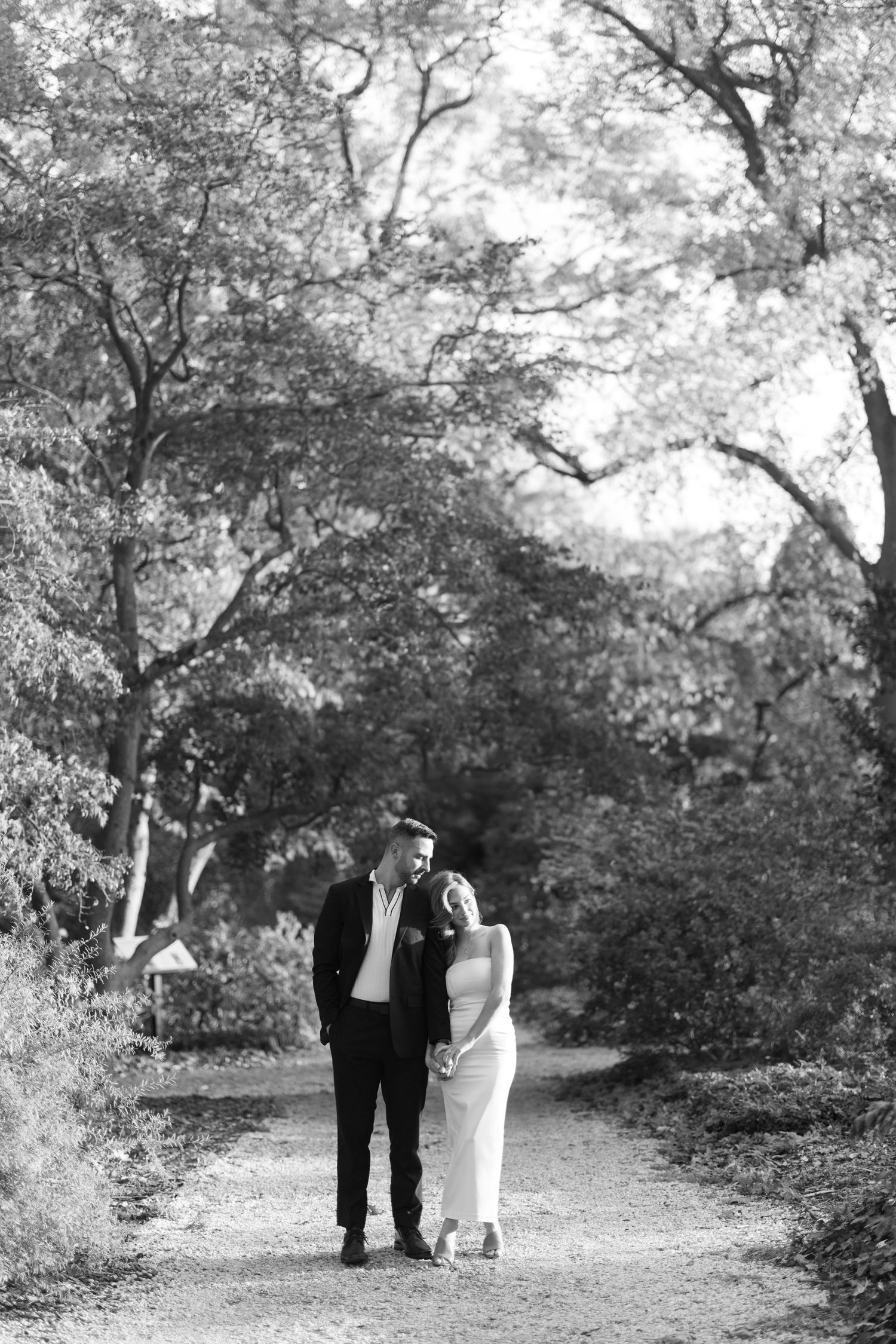Black and white photo of a couple walking hand in hand along a tree-lined garden path in elegant attire.
