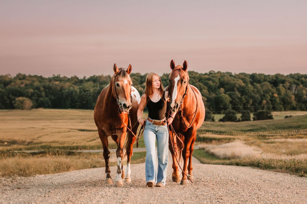 Woman in jeans walking with two brown horses on dirt road during sunset.