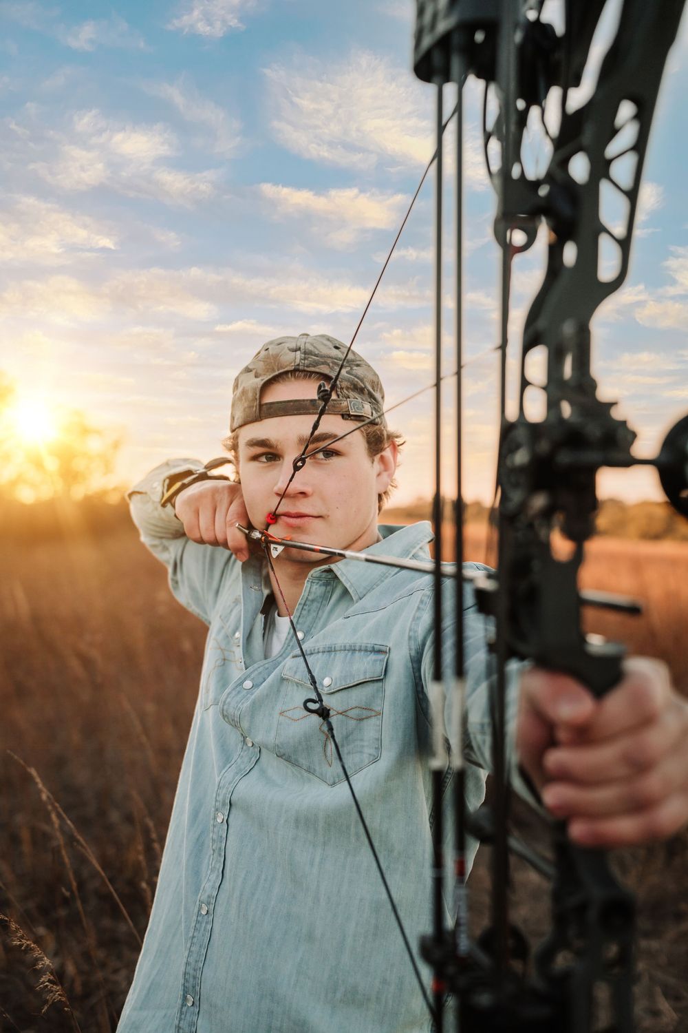 Archer in camouflage cap draws compound bow at sunset against golden sky with silhouetted archery equipment.