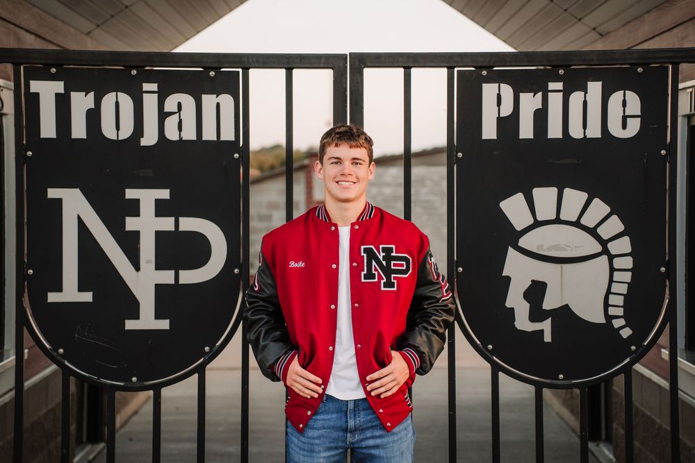A varsity letterman stands between two large black circular Trojan Pride logos mounted on a wall.