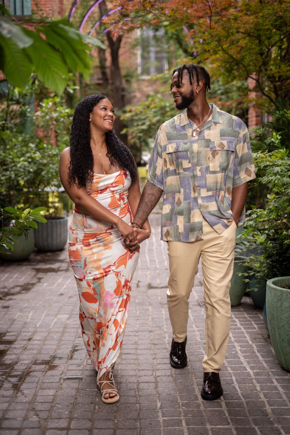 A happy Black couple walks hand in hand along a charming brick garden path lined with lush plants.