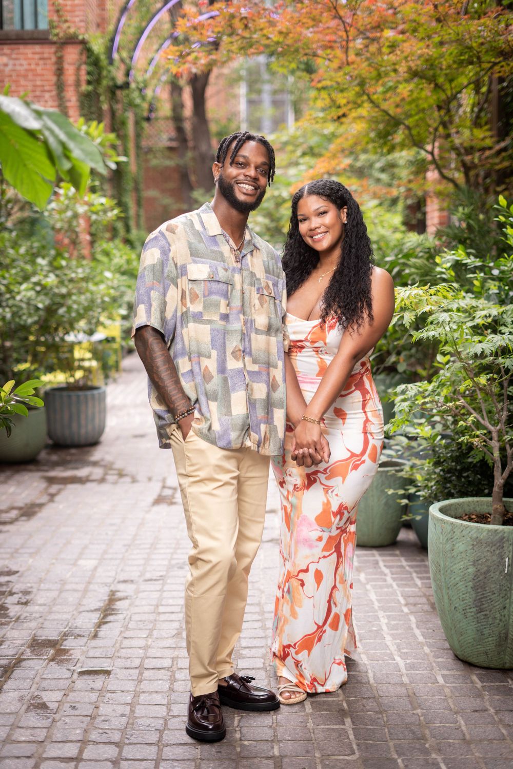 A stylish Black couple holds hands and poses together on a brick garden pathway surrounded by greenery.