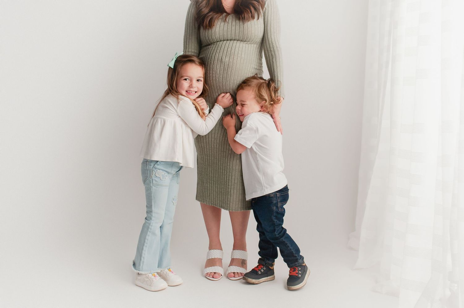 Two young children hugging pregnant mothers belly in bright white studio portrait.