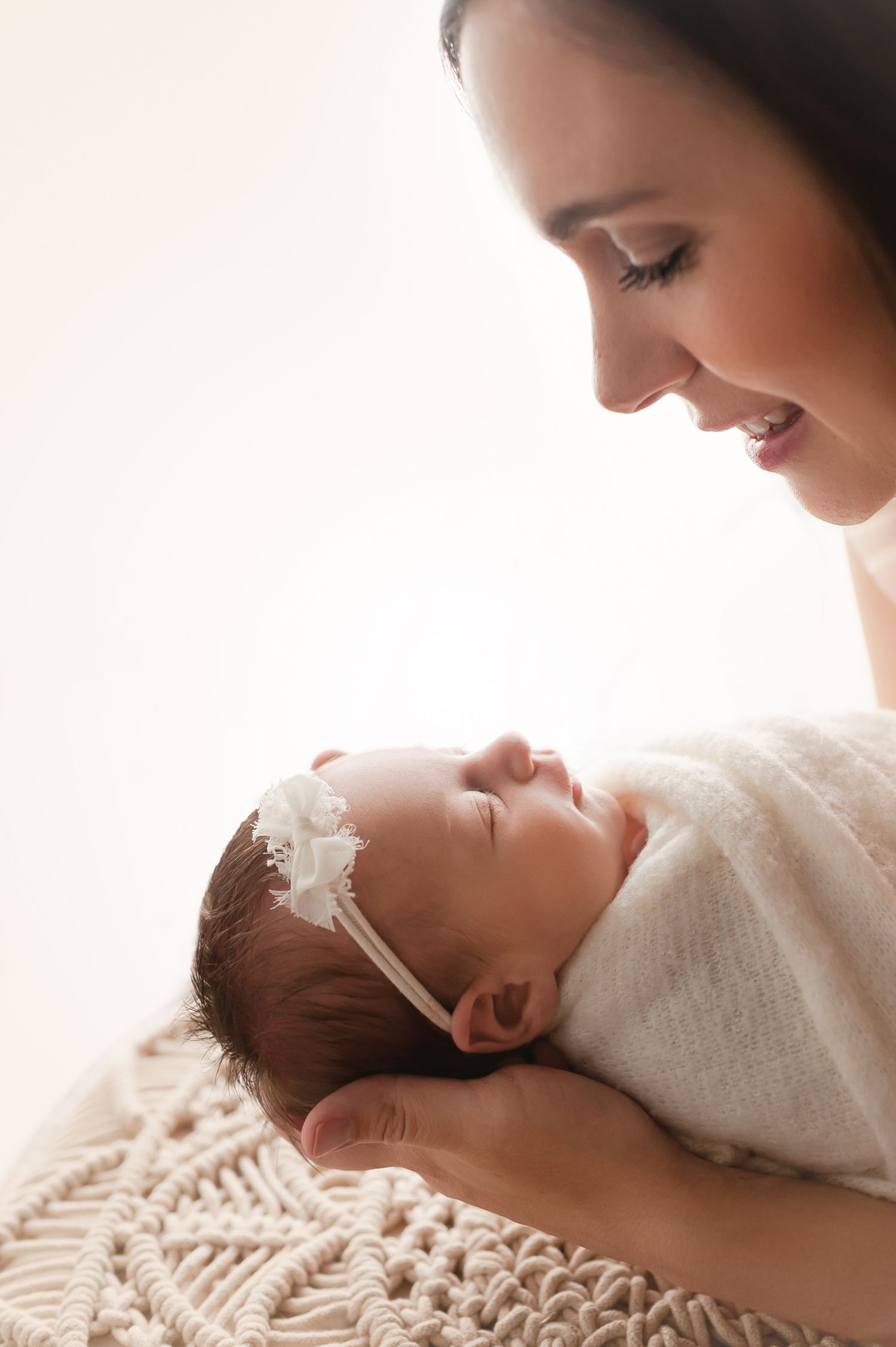 A smiling mother gazes lovingly at her sleeping newborn baby girl wrapped in white with a floral headband.
