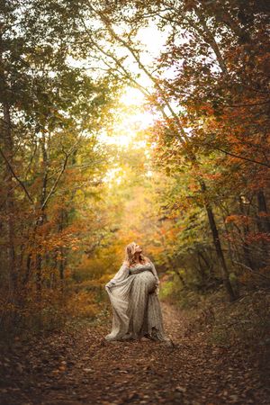 A pregnant woman poses gracefully along a leaf-covered forest path during golden hour among autumn trees for maternity photos.