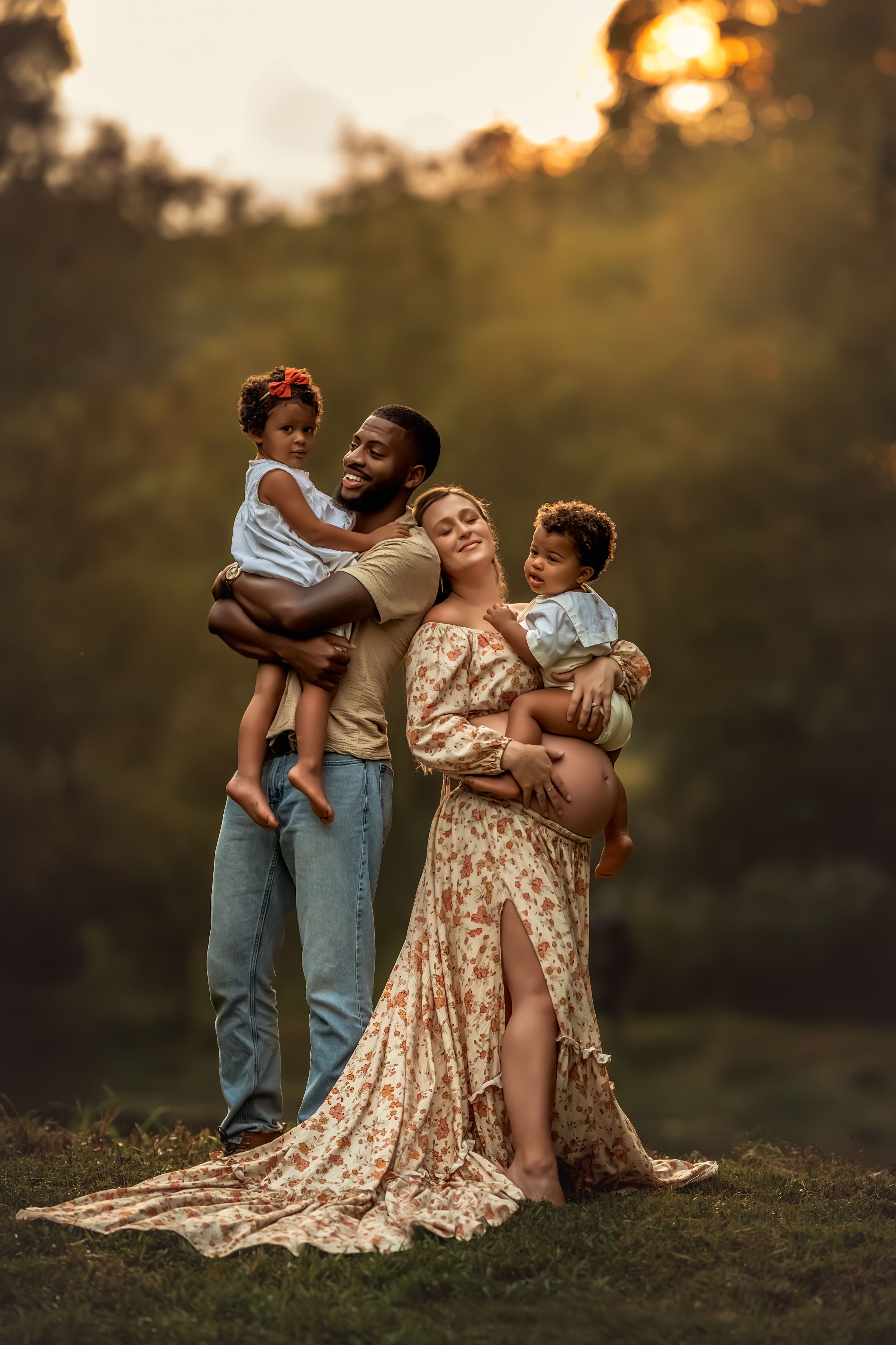 A joyful interracial family poses outdoors at golden hour, with a pregnant mother and father each holding a young child for maternity photos