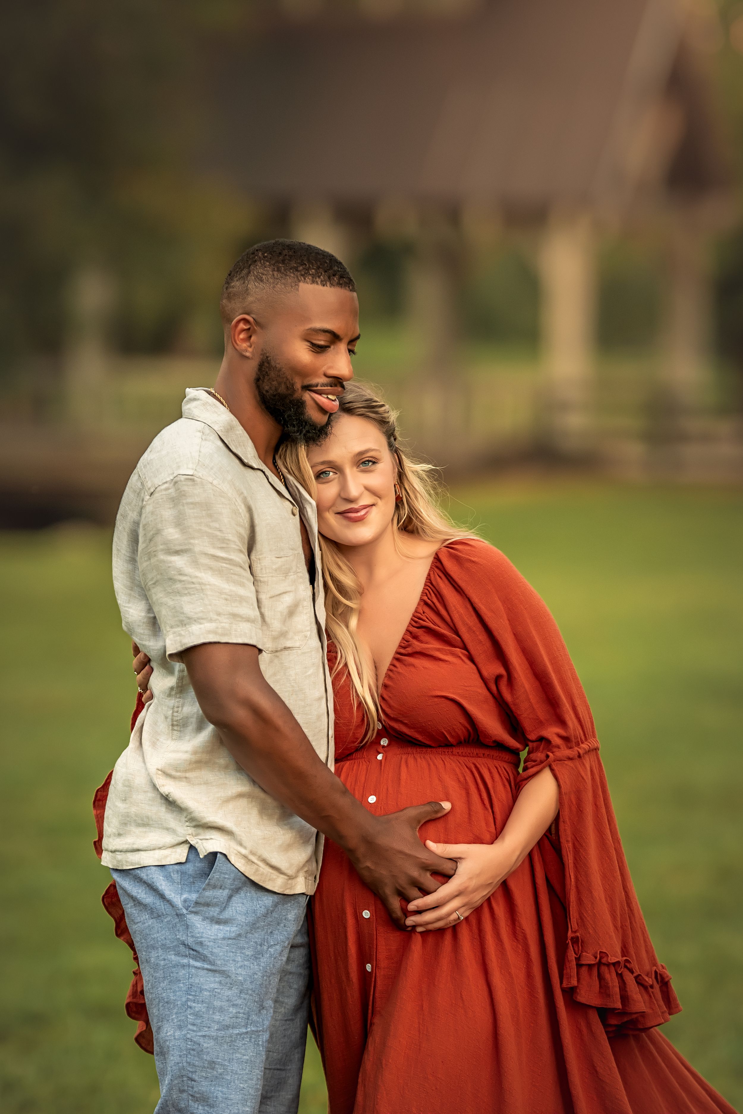 Interracial couple sharing a tender maternity photo outdoors in North Georgia, woman in rust color dress cradling baby bump.