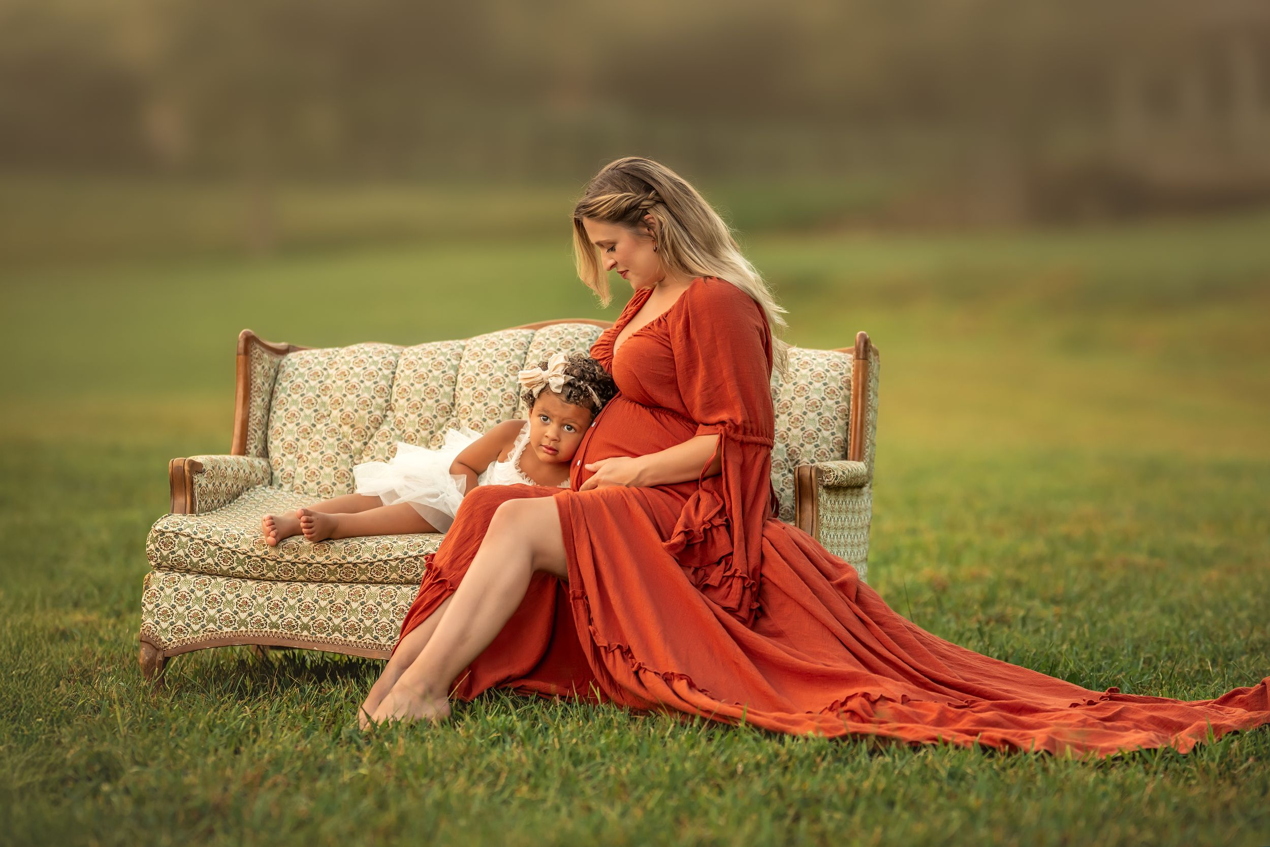 Pregnant woman in rust dress sits on vintage sofa outdoors with young child on her lap in a lush green field in North Georgia.