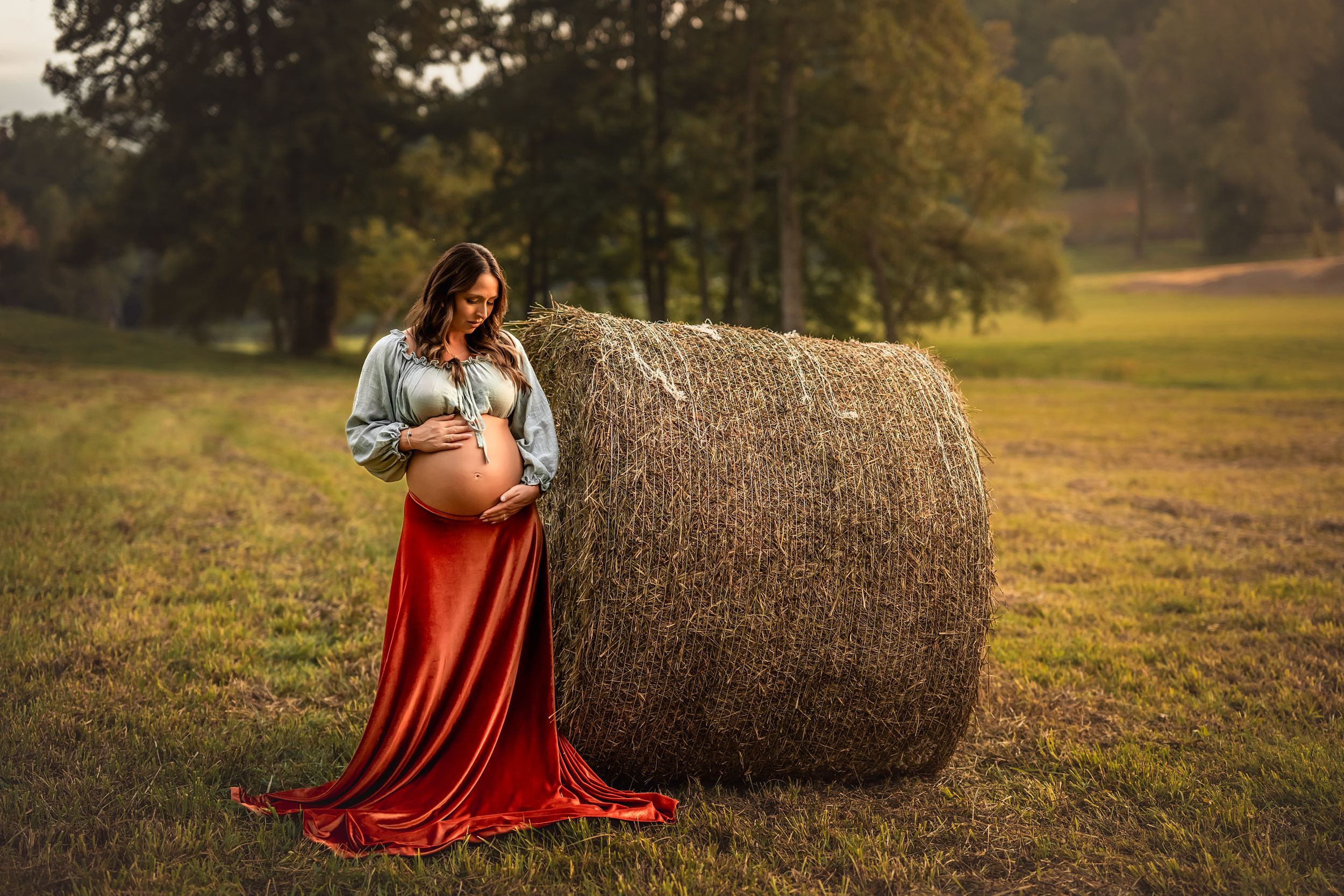 Pregnant woman in velvet skirt and denim crop top poses with bare belly beside a hay bale in a golden sunlit field.
