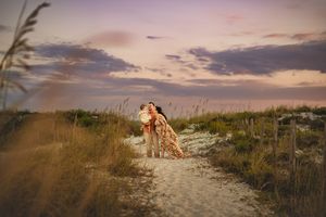A pregnant mom and dad embraces toddler on a sandy beach path at sunset with sea oats swaying in the foreground and pink clouds overhead.
