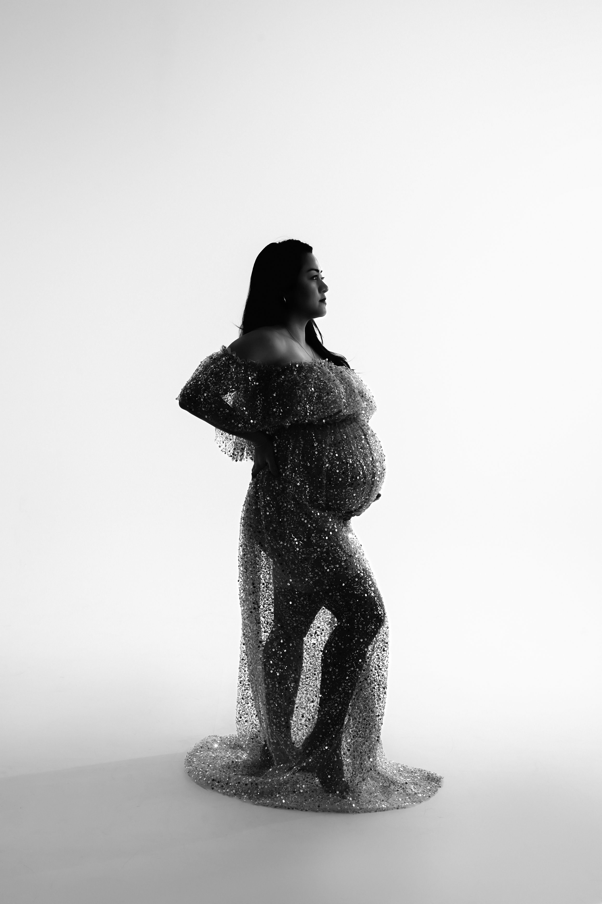 A black and white backlit studio photo of pregnant woman in sheer sparkly dress for fine art maternity photos in studio.