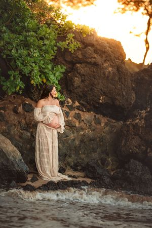 Woman in a flowing beige dress poses for bare belly maternity photos on beach against dramatic volcanic rocks and foliage at sunset.