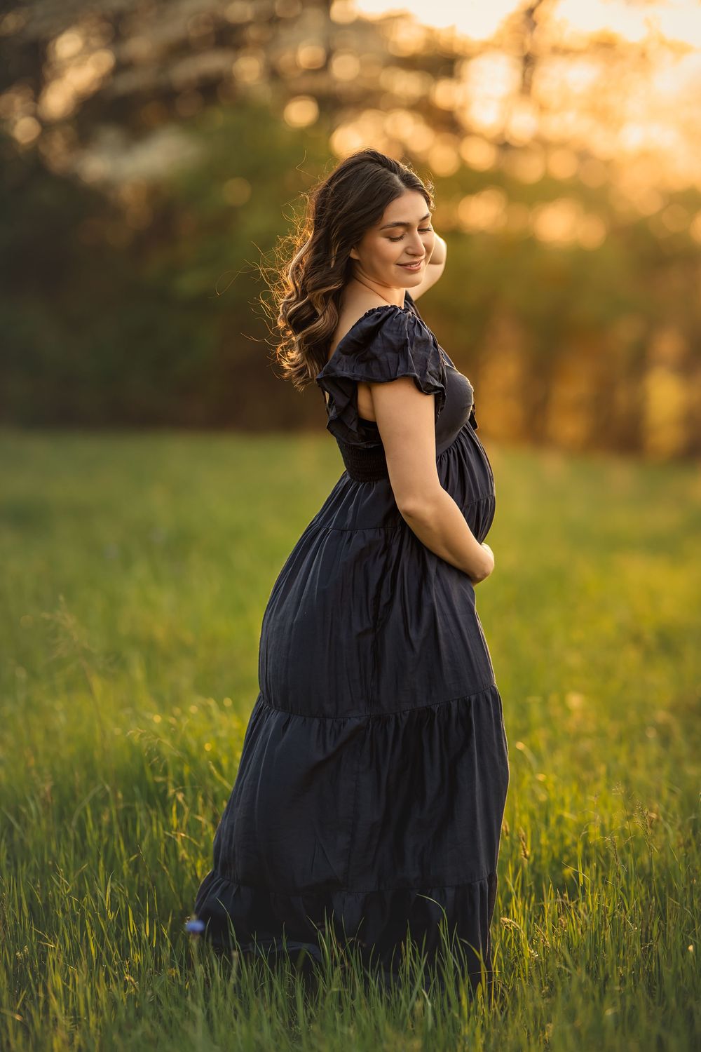 A pregnant mother in a flowing navy dress stands in a sunlit meadow during golden hour with windswept hair.