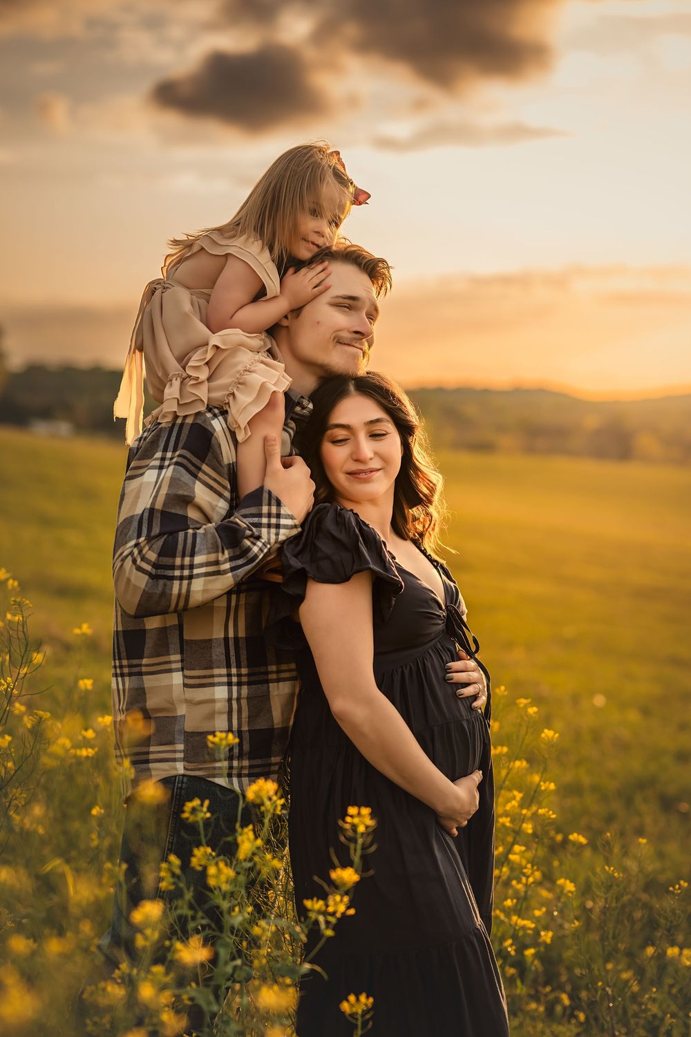 Dad holds toddler on shoulders while hugging pregnant wife in a golden field at sunset as warm light bathes the pastoral landscape.