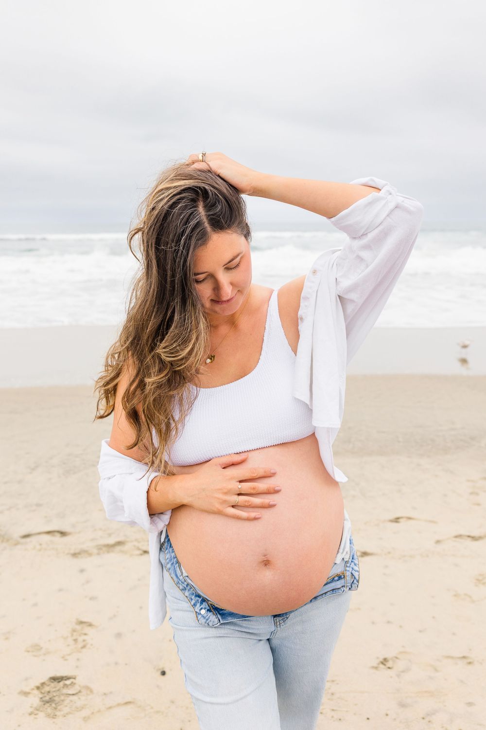 Expectant mother in white tank top and jeans stands on beach touching her pregnant belly on cloudy day.