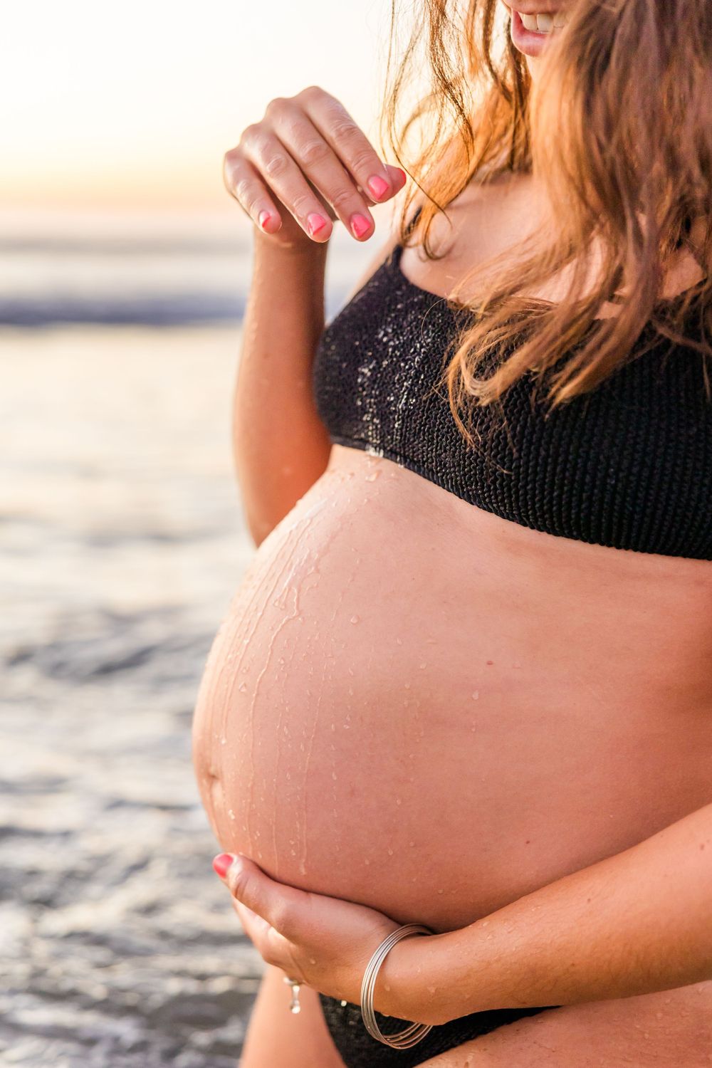 Pregnant person in a black swimsuit holds their belly while standing on a beach at sunset. carlsbad maternity photographer