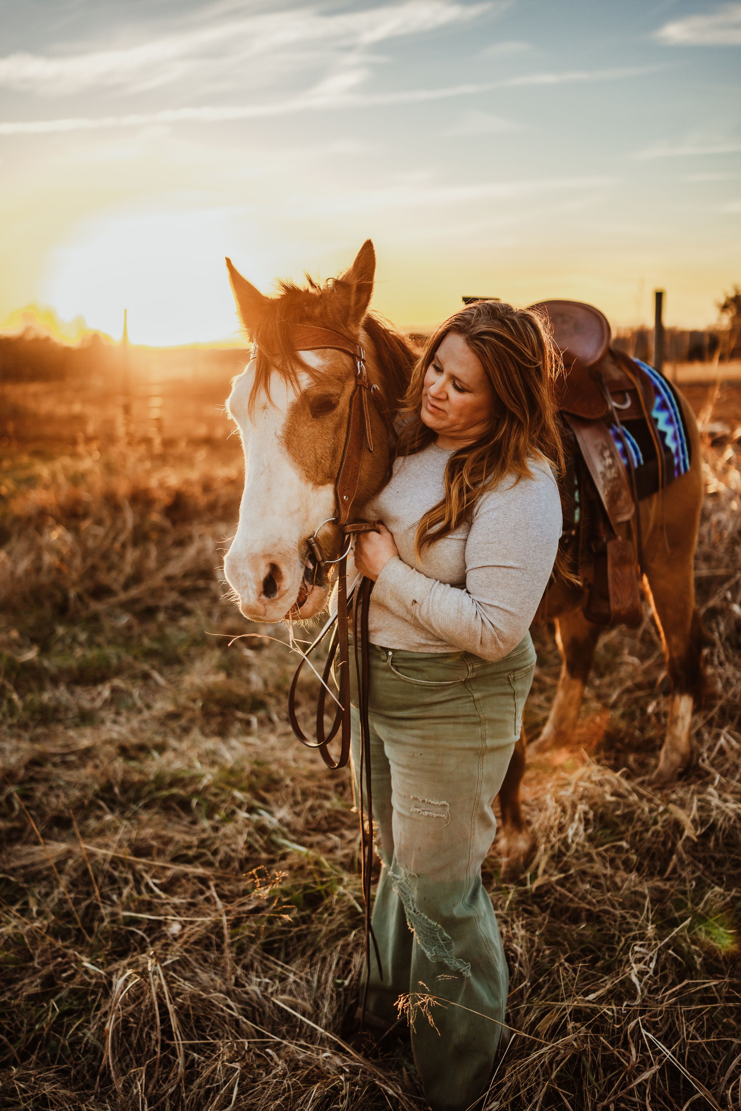 A person and two horses stand together in a golden sunlit field during sunset, creating a warm and peaceful scene.
