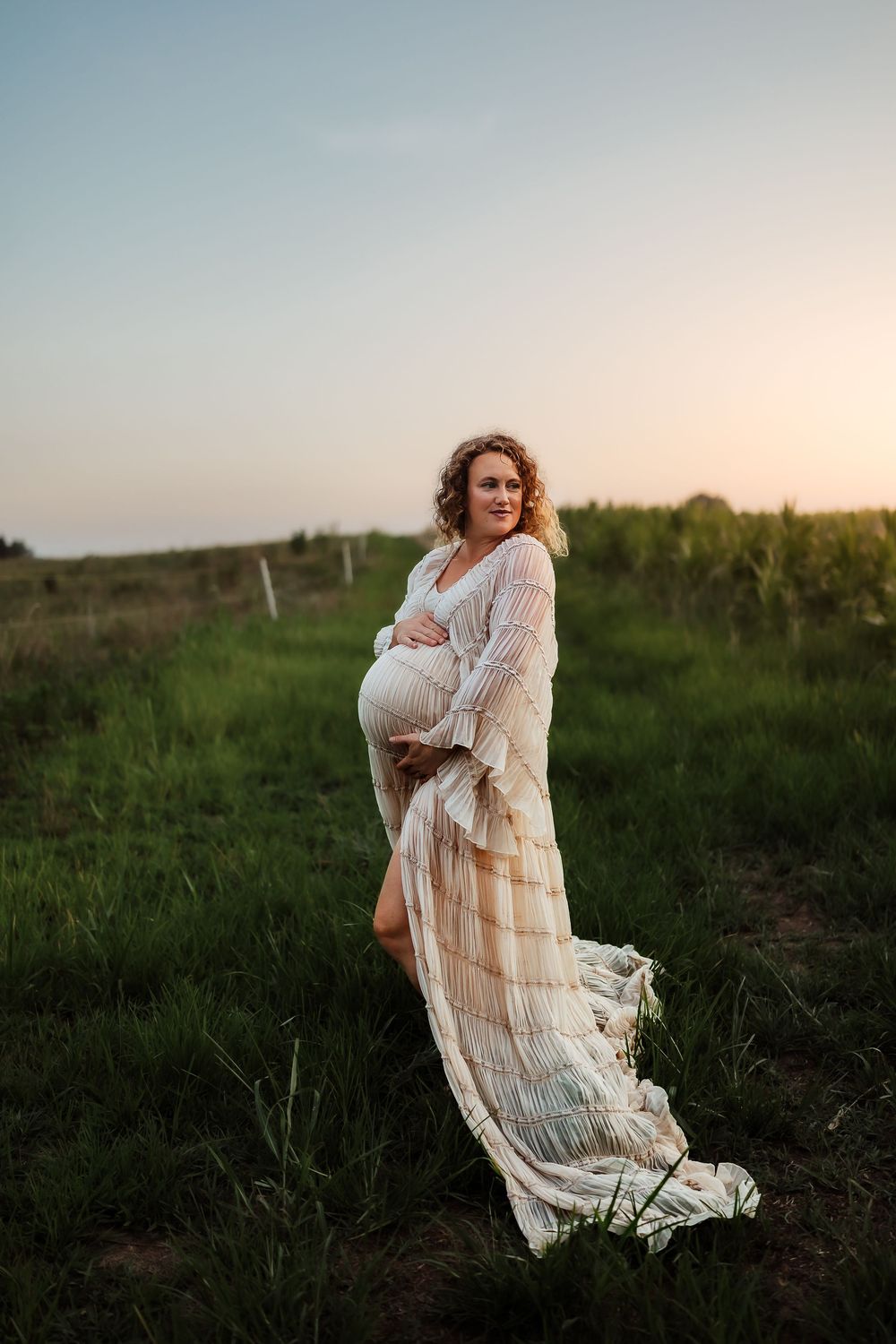 Pregnant woman in flowing white dress cradling her belly in a green field at golden hour sunset.