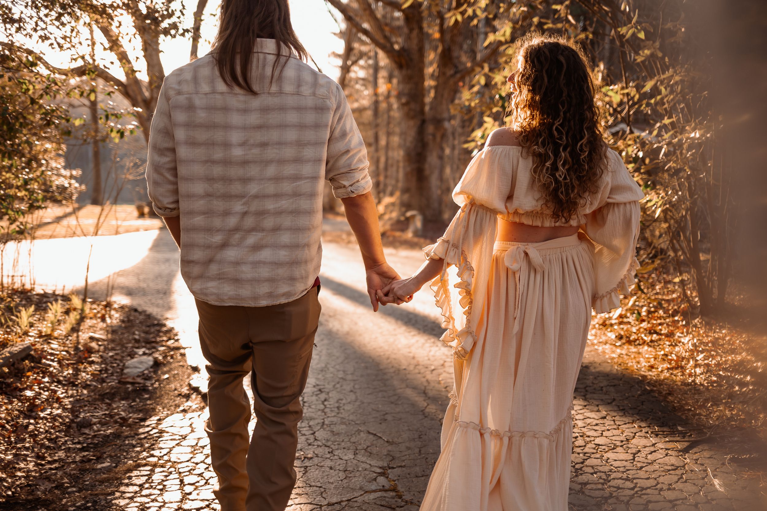 A couple holding hands while walking along a sunlit path through autumn trees.