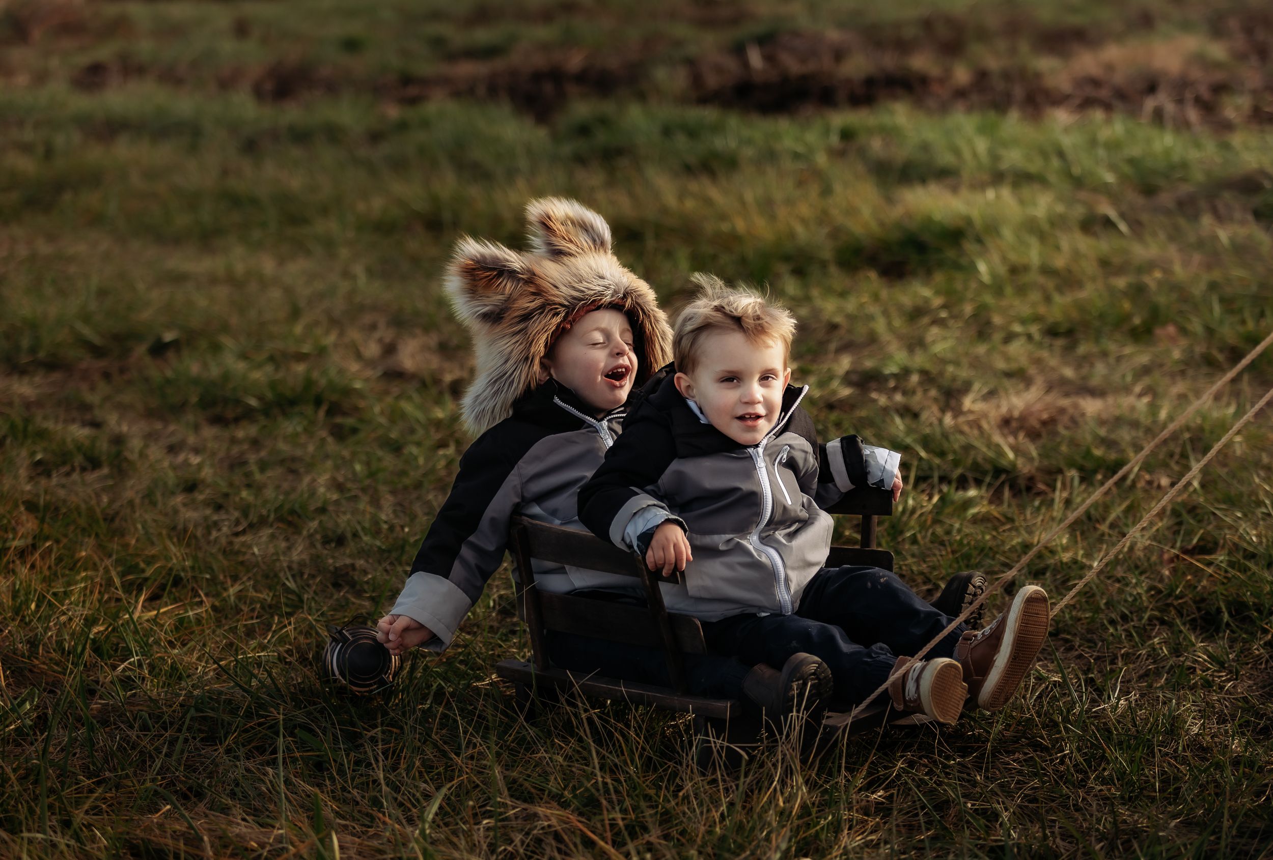 Two children laugh while riding in wooden wagon through grassy field.