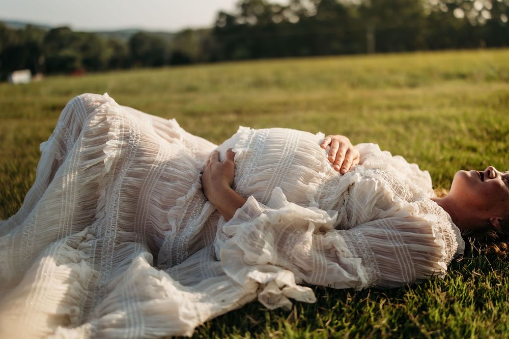 A pregnant woman in a white lace dress lies peacefully on green grass in a sunlit field.
