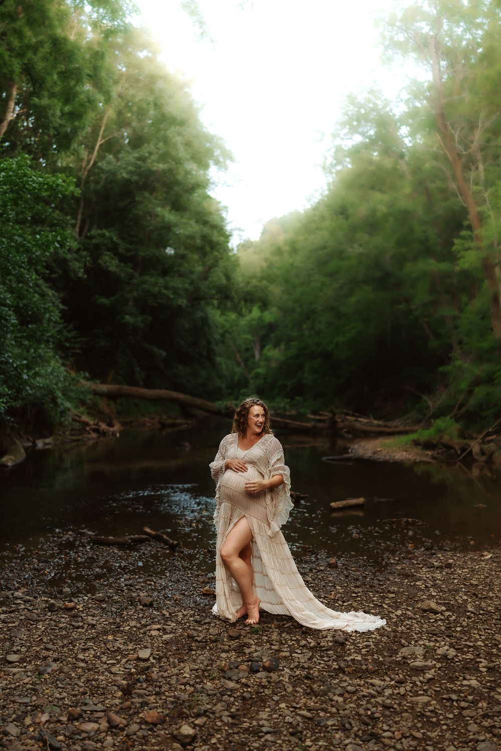 Pregnant woman in white lace gown standing in a rocky creek surrounded by lush green forest trees.