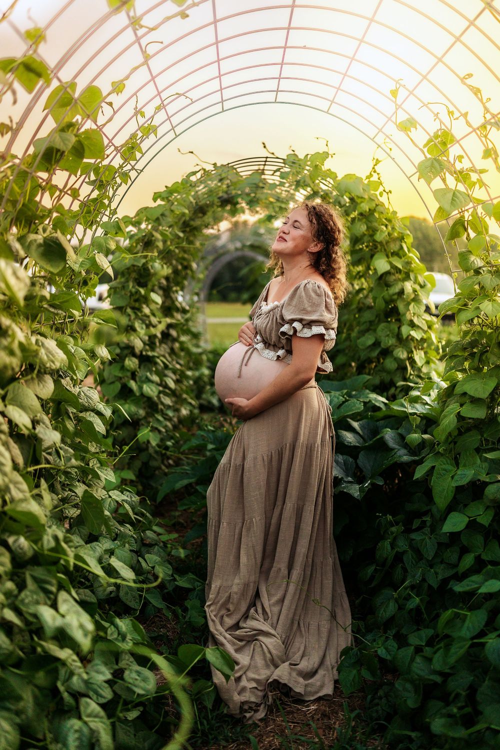 Pregnant woman in olive dress cradling belly in lush green garden tunnel at golden hour.