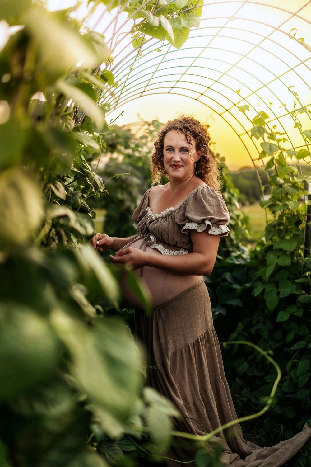 A curly-haired woman in a brown dress tends to lush green plants inside a greenhouse at golden hour.