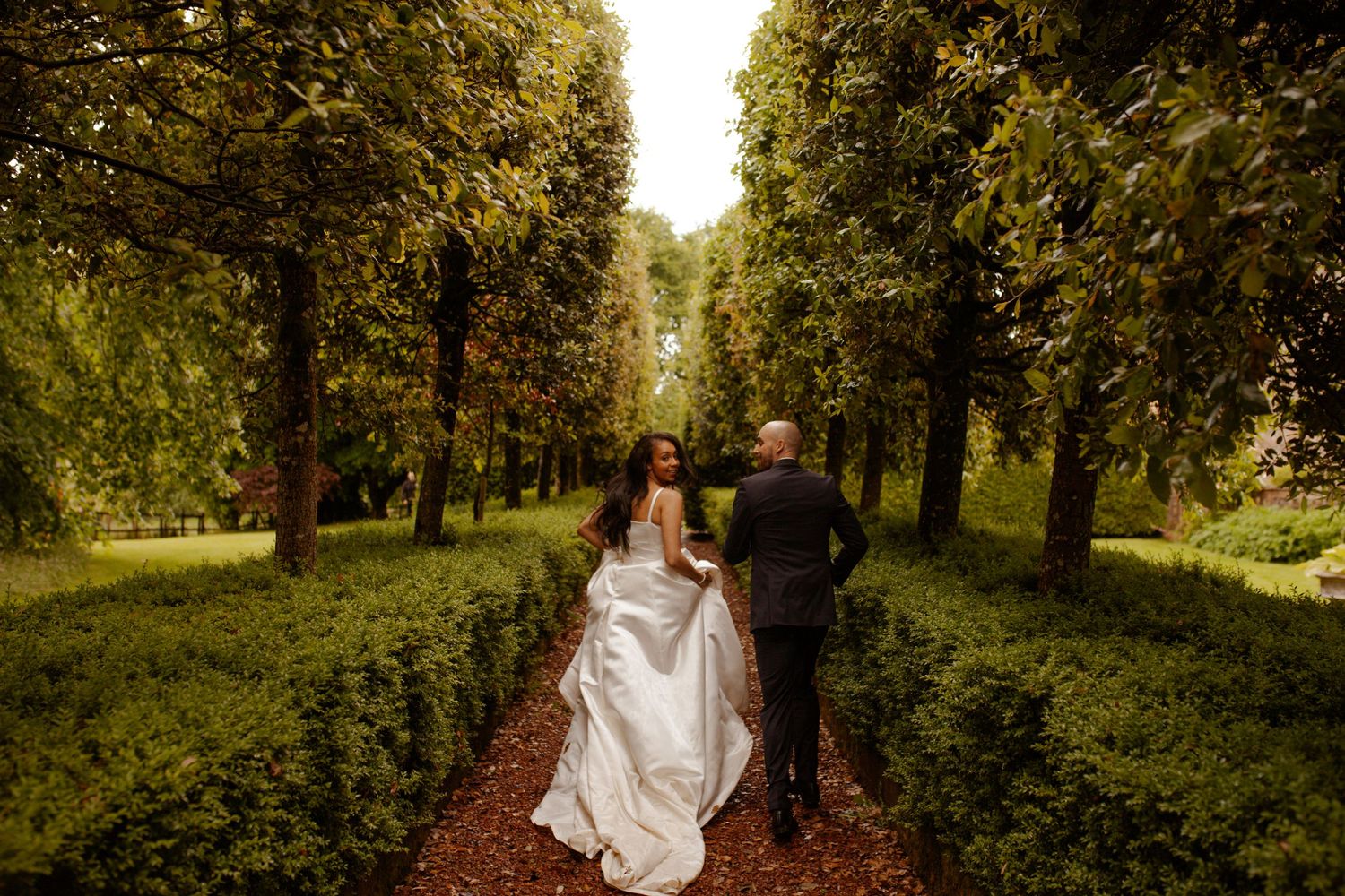 A couple walks hand in hand through a scenic tree-lined path with golden sunlight filtering through.