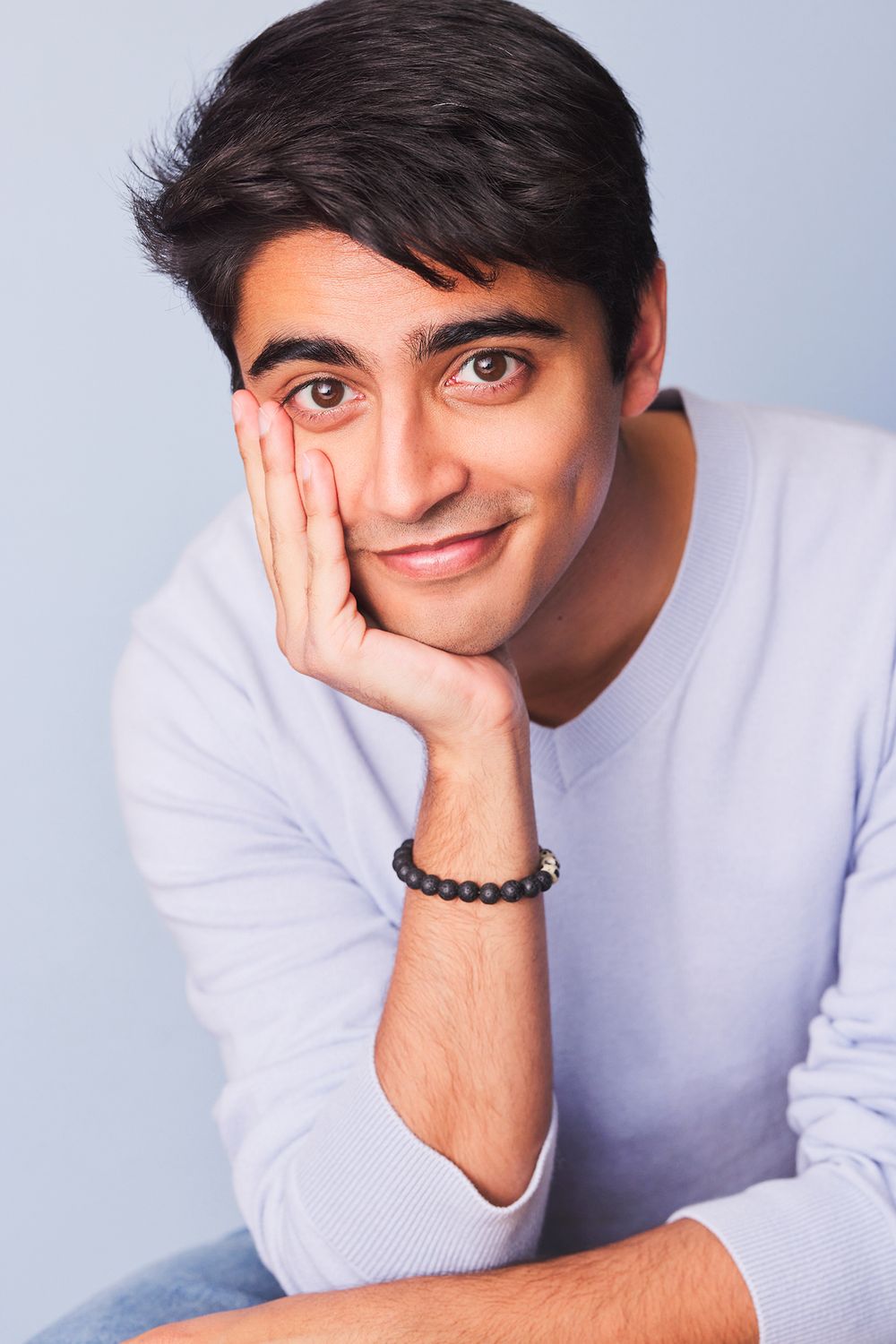 Actor in a white sweater poses playfully with hands on their face against a light gray background.