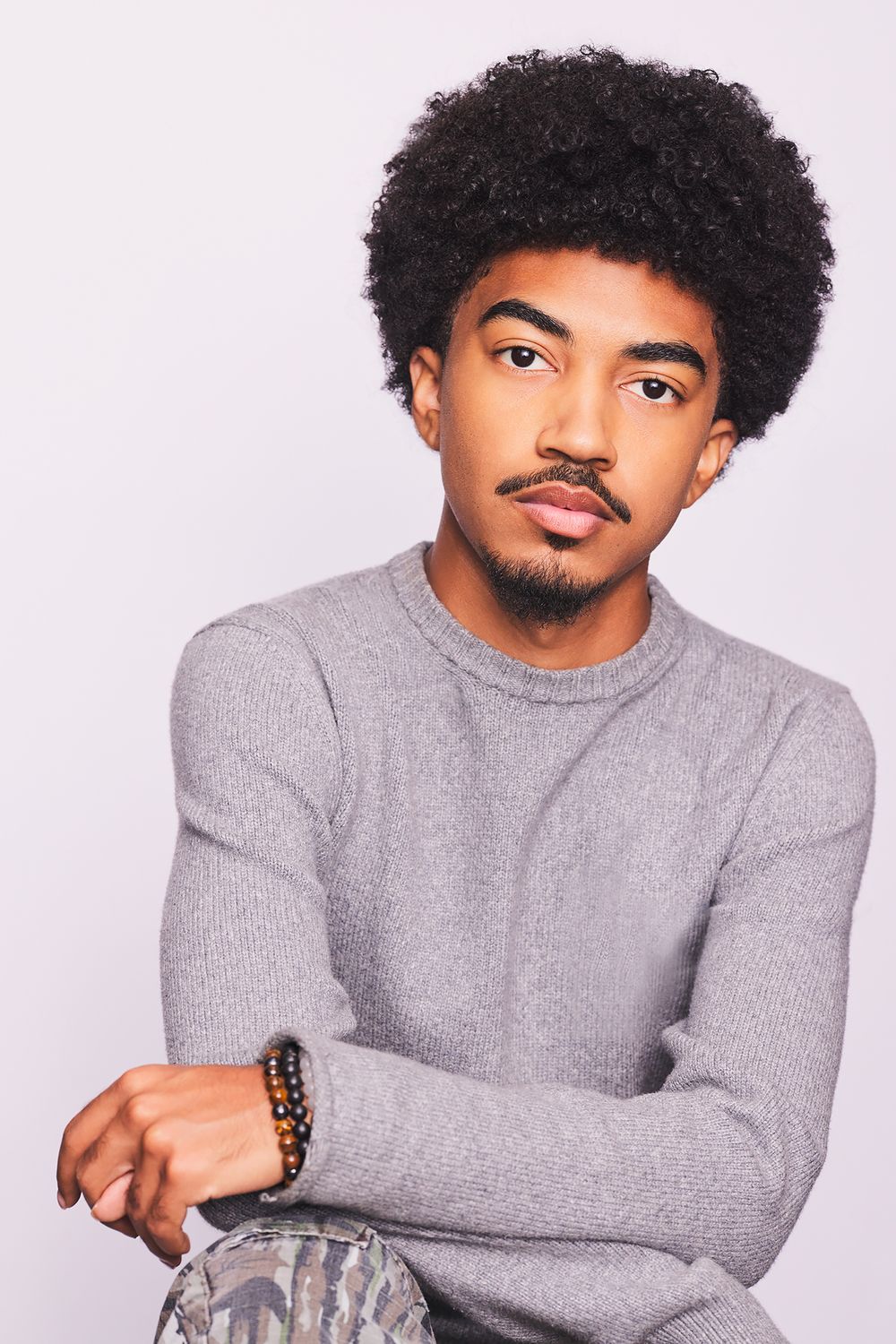 Theatrical Headshots of a person of color with curly hair wearing a gray sweater posing against a light background.