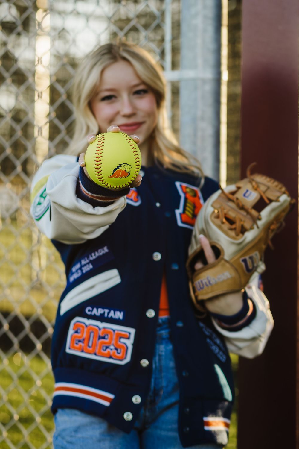 A softball player holding up a yellow ball while wearing a varsity jacket and glove.