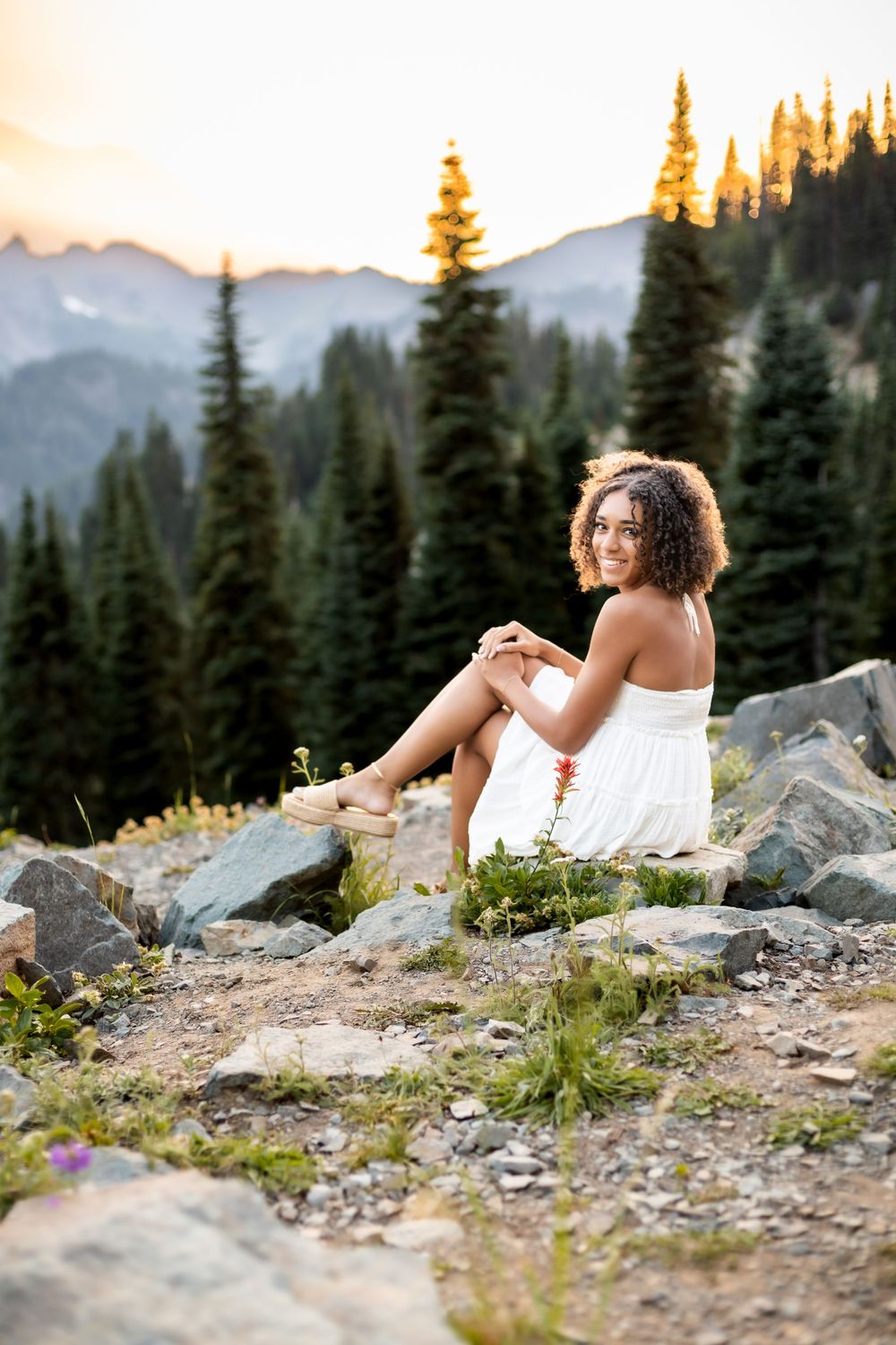 A scenic mountain portrait at sunset wearing a white dress among evergreen trees.