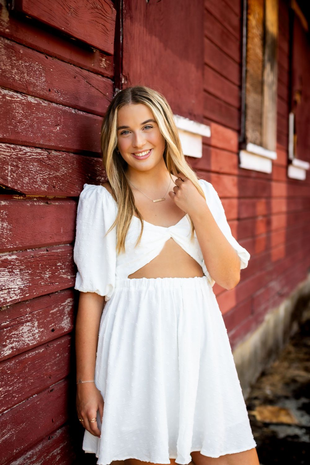 A white cutout dress is modeled against a rustic red barn wall in warm lighting.