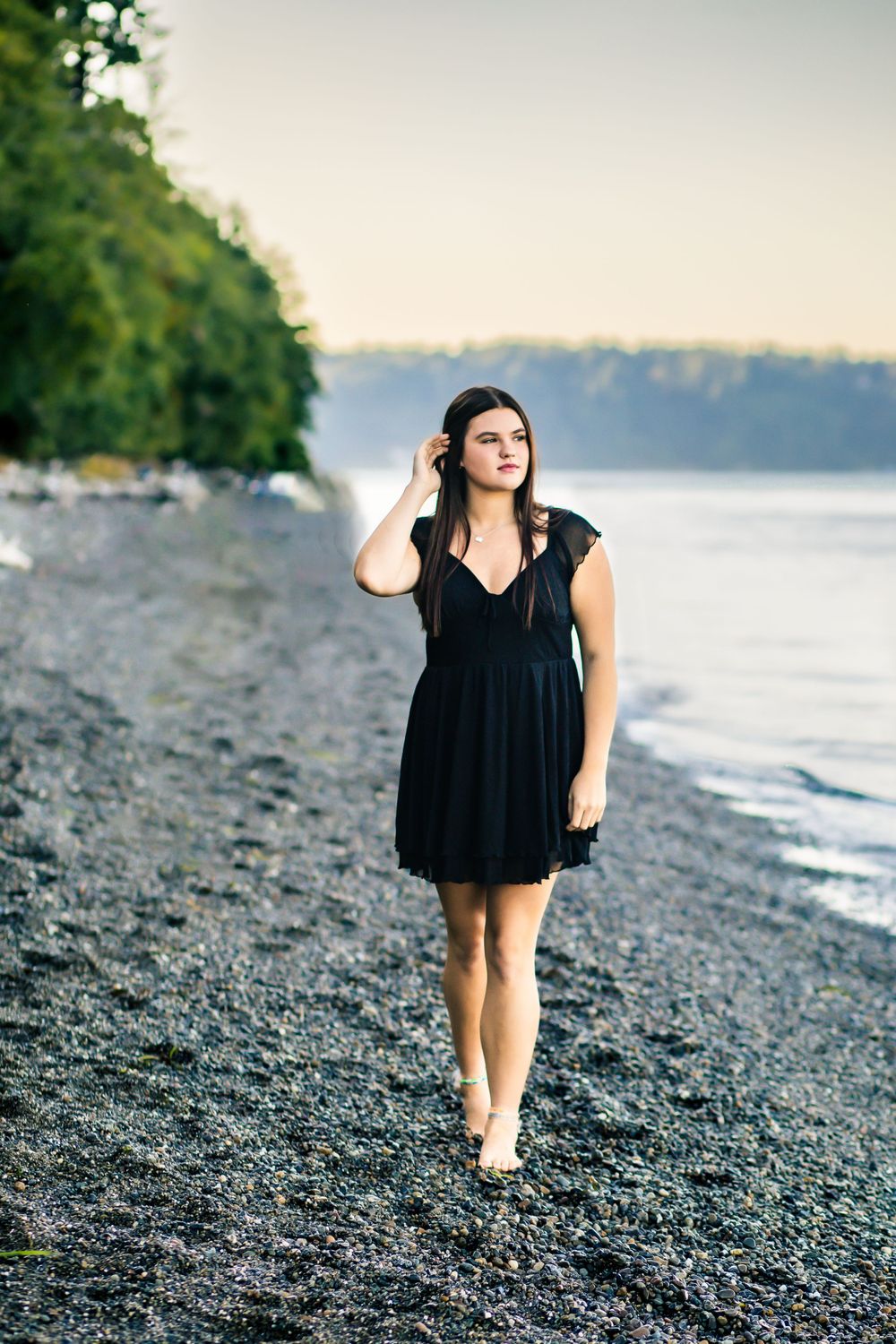 A woman in a black dress stands thoughtfully on a rocky beach at sunset.