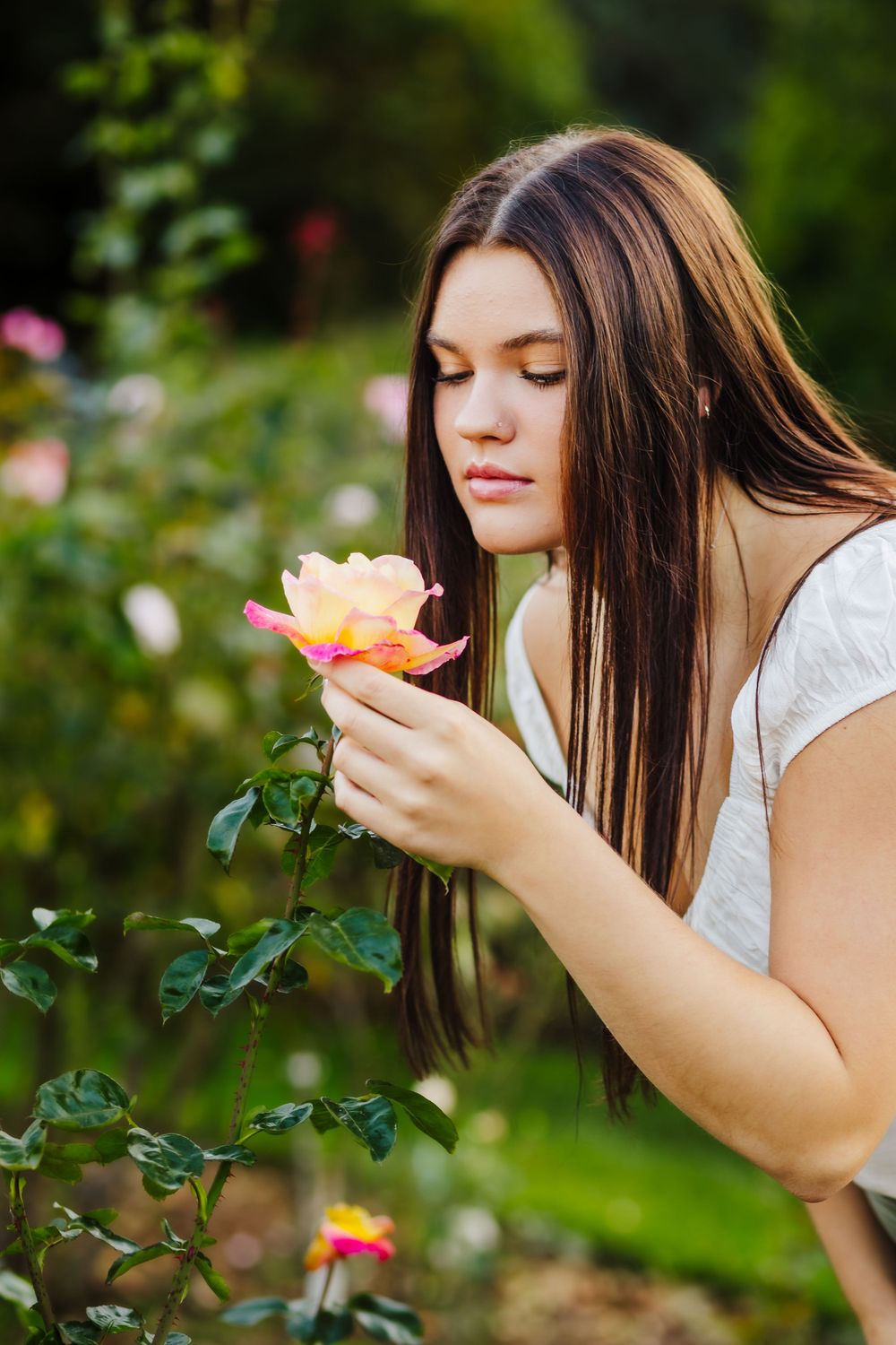 A woman admires and smells a peachy-orange rose in a lush garden setting.