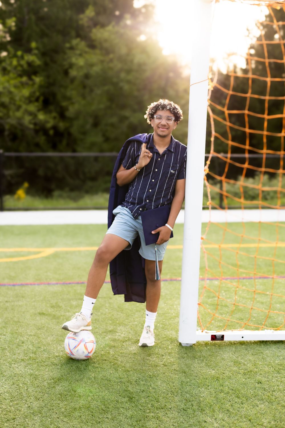 Soccer player crouching near goal post on green grass field at sunset with orange netting in background.