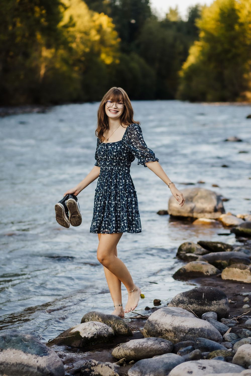 A flowing blue dress is worn while standing on river rocks in a scenic outdoor setting.